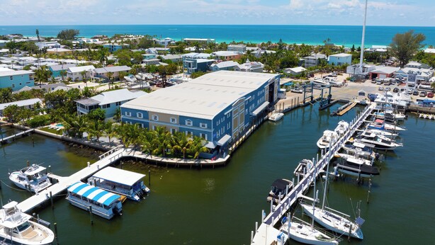 A marina with multiple boats docked, a large building with a blue roof, and a clear view of the ocean. - AMI Condo W/Pool, Walk to Beach, Bars, Restaurants and Fishing! (Bradenton Beach)