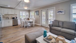A spacious living area with a gray sofa, a dining table with chairs, a kitchen with white cabinets, and a ceiling fan. - AMI Condo W/Pool, Walk to Beach, Bars, Restaurants and Fishing! (Bradenton Beach)