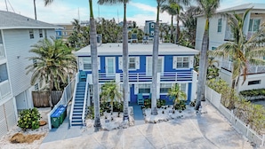 A two-story building with a blue and white exterior, a front entrance with a staircase, and a small front yard with palm trees and a white picket fence. - AMI Condo W/Pool, Walk to Beach, Bars, Restaurants and Fishing! (Bradenton Beach)
