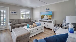 A living room with a sofa, ottoman, TV, and a wall-mounted artwork labeled "beach". - AMI Condo W/Pool, Walk to Beach, Bars, Restaurants and Fishing! (Bradenton Beach)