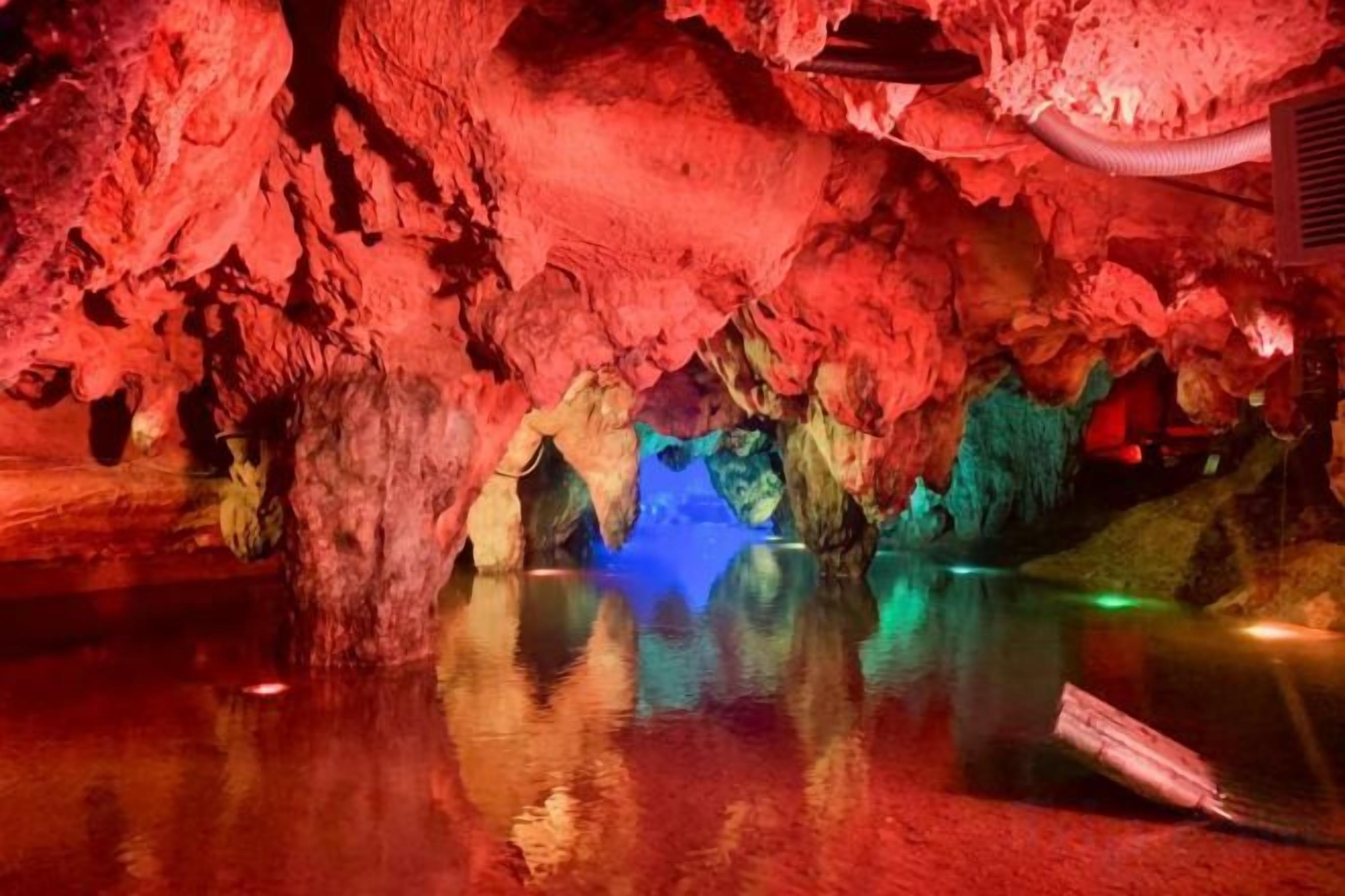 Eine Höhle mit Stalaktiten und Stalagmiten, beleuchtet durch rote und blaue Lichter, und eine spiegelnde Wasseroberfläche.