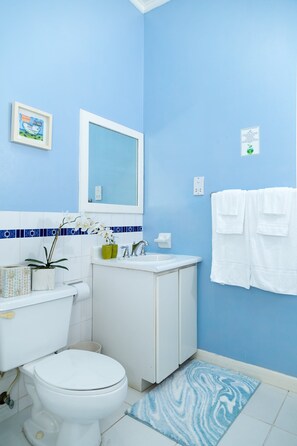 A bathroom with a toilet, sink, and mirror. There are towels hanging on the wall and a small potted plant on the counter. - 🏝️ Sleep to Caribbean Waves Pool | Beach + Pool + AC • Ocho Rio (Ocho Rios)