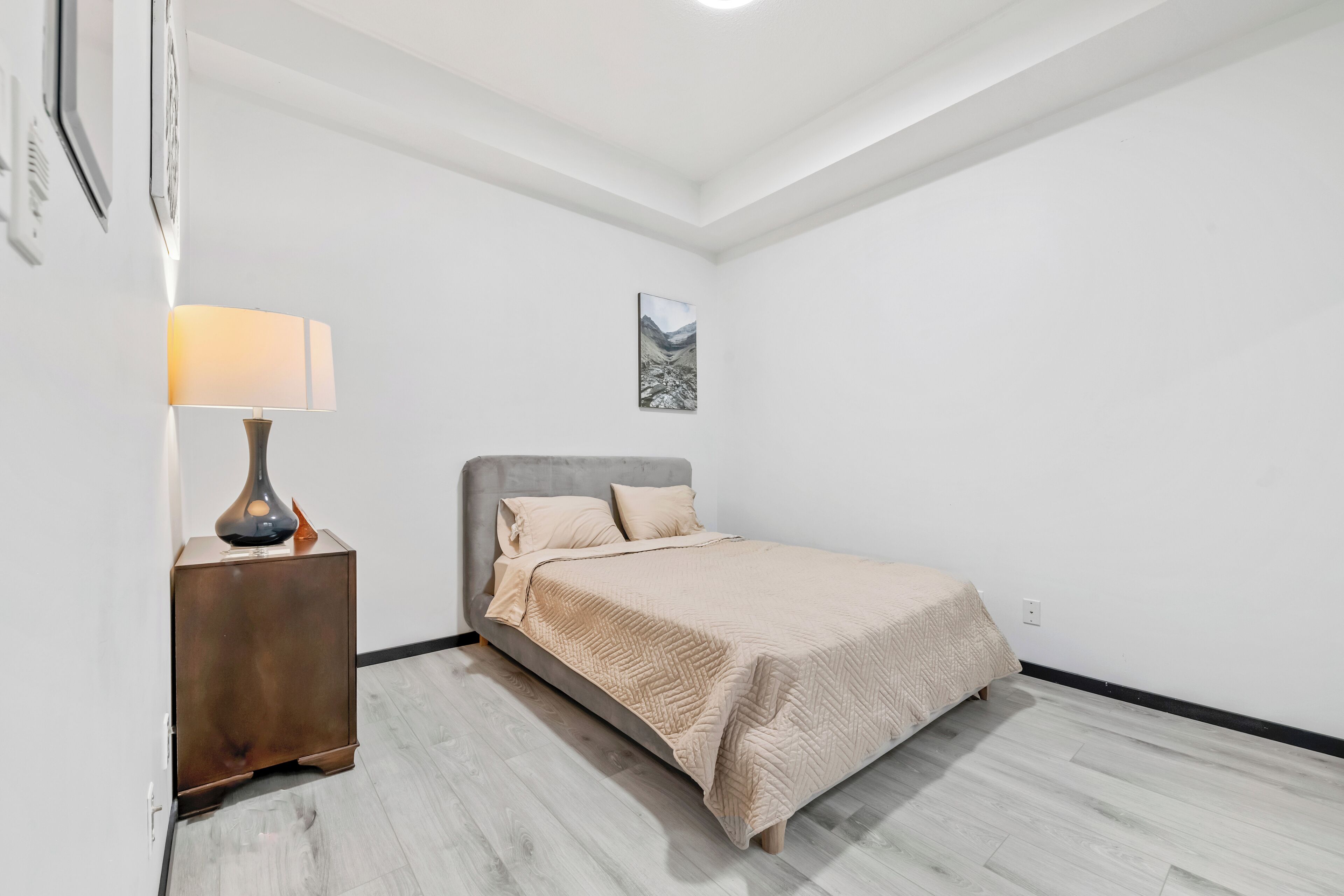 Un dormitorio con cama, mesita de noche, lámpara y un cuadro en la pared.