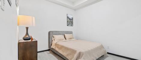 Un dormitorio con cama, mesita de noche, lámpara y un cuadro en la pared.