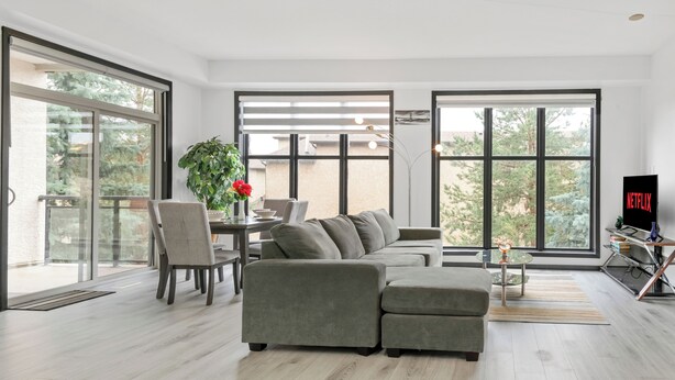Uma sala de estar moderna com um sofá seccional, uma área de jantar com mesa e cadeiras, e uma janela grande com vista para árvores.