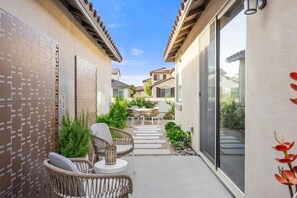 Un patio exterior estrecho con mobiliario de mimbre, una pequeña mesa y un ventanal de vidrio que da a un jardín.