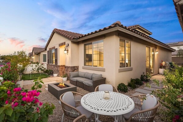 A residential building with a patio area featuring a round table and chairs, a built-in fire pit, and a grey sofa.