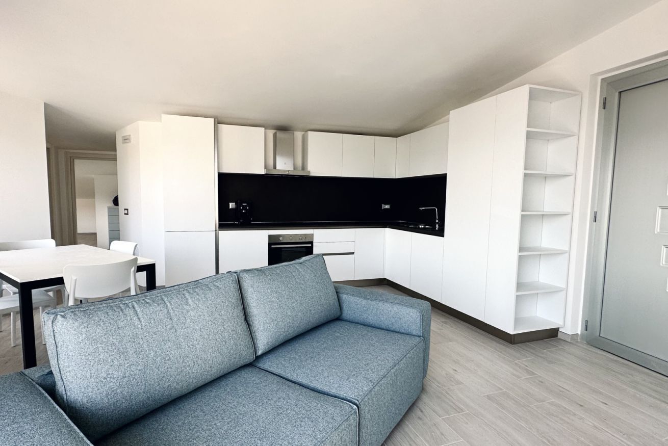 Un soggiorno moderno con un divano grigio, una cucina con mobili bianchi e un'area pranzo con tavolo e sedie bianche.