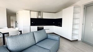 Un saló modern amb un sofà gris, una cuina amb armaris blancs i una zona de menjador amb taula i cadires blanques.
