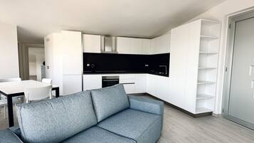 Un soggiorno moderno con un divano grigio, una cucina con mobili bianchi e un'area pranzo con tavolo e sedie bianche.