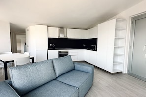 Un salon moderne avec un canapé gris, une cuisine avec des meubles blancs et un coin repas avec une table et des chaises blanches.