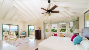 Une chambre spacieuse avec un grand lit, un ventilateur de plafond et une vue sur l’océan depuis les portes du balcon.