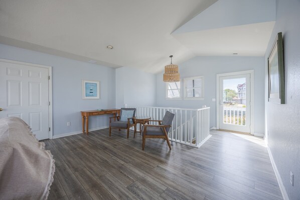 A room with a wooden desk, two chairs, a hanging light fixture, and a view through a door.