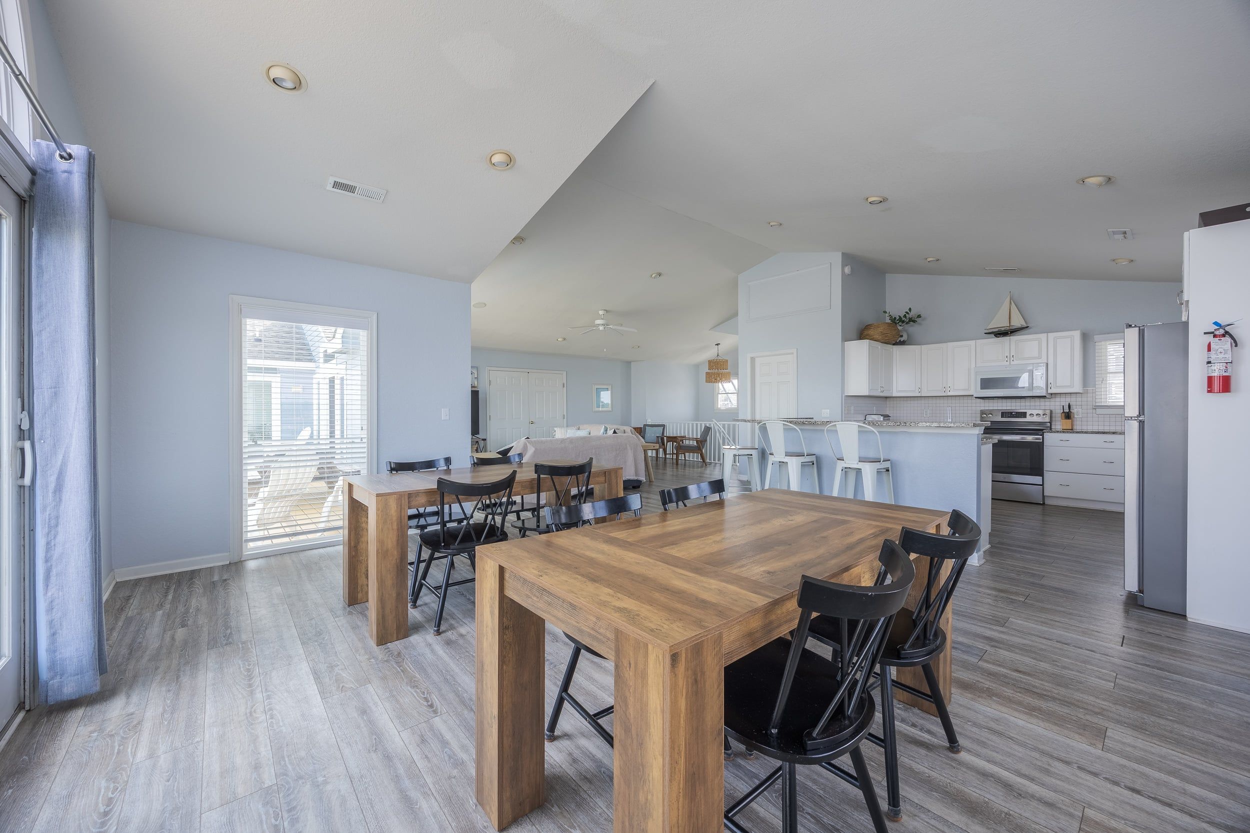 A spacious living area with a large wooden dining table, a kitchen with white cabinets, and a view through a large window.