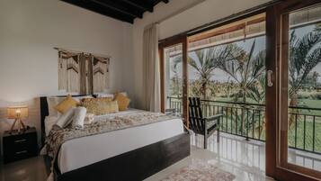 A bedroom with a bed, a nightstand, a lamp, a window with a view of palm trees, and a balcony with a chair.