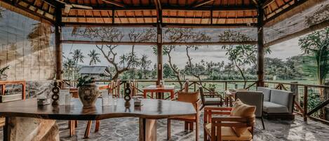 An outdoor dining area with a thatched roof, stone flooring, and a view of palm trees and hills.