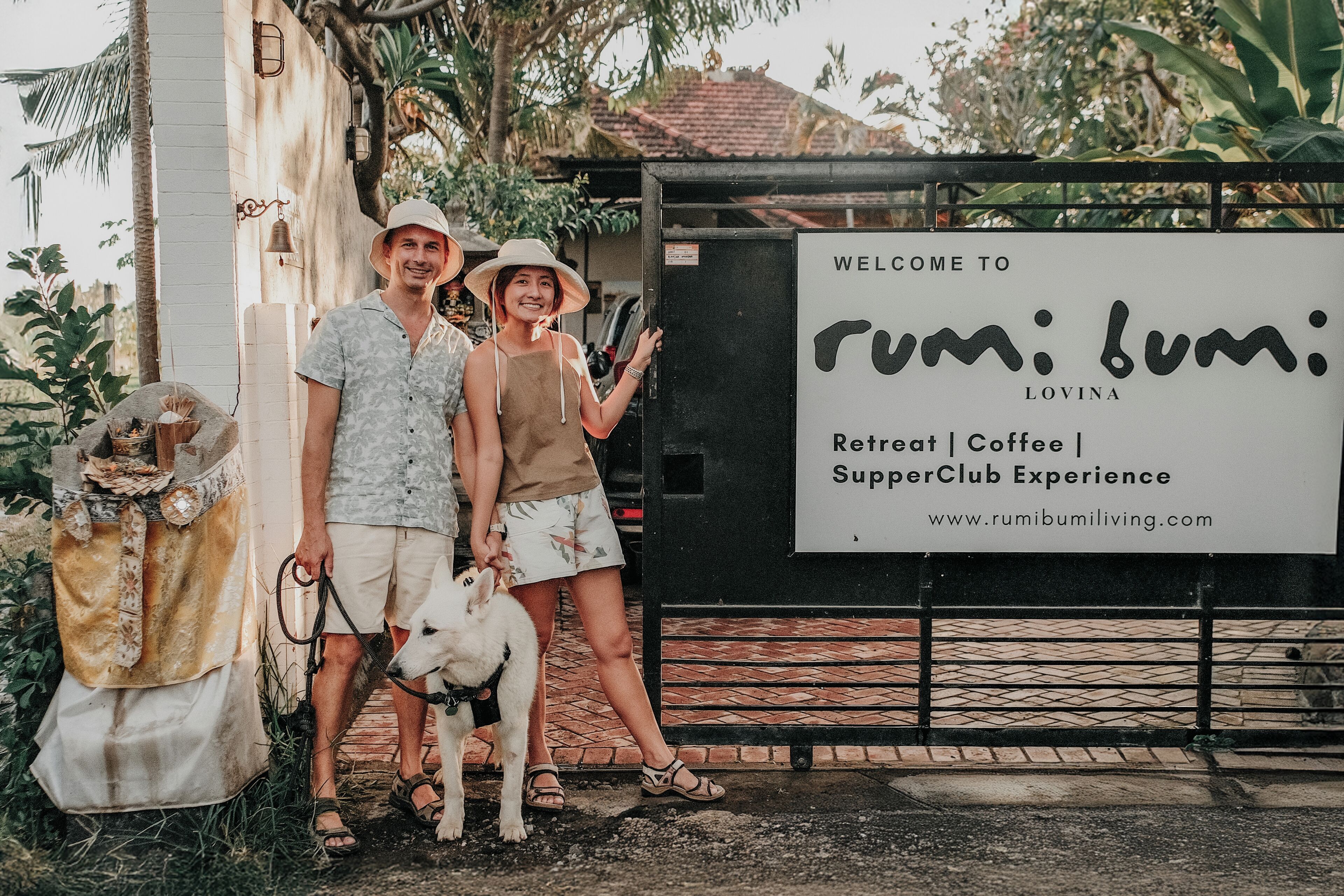 Un couple, accompagné de son chien, se tient devant un panneau indiquant : « Bienvenue sur Rumilumiliving.com, lieu de retraite | café | expérience SupperClub ».