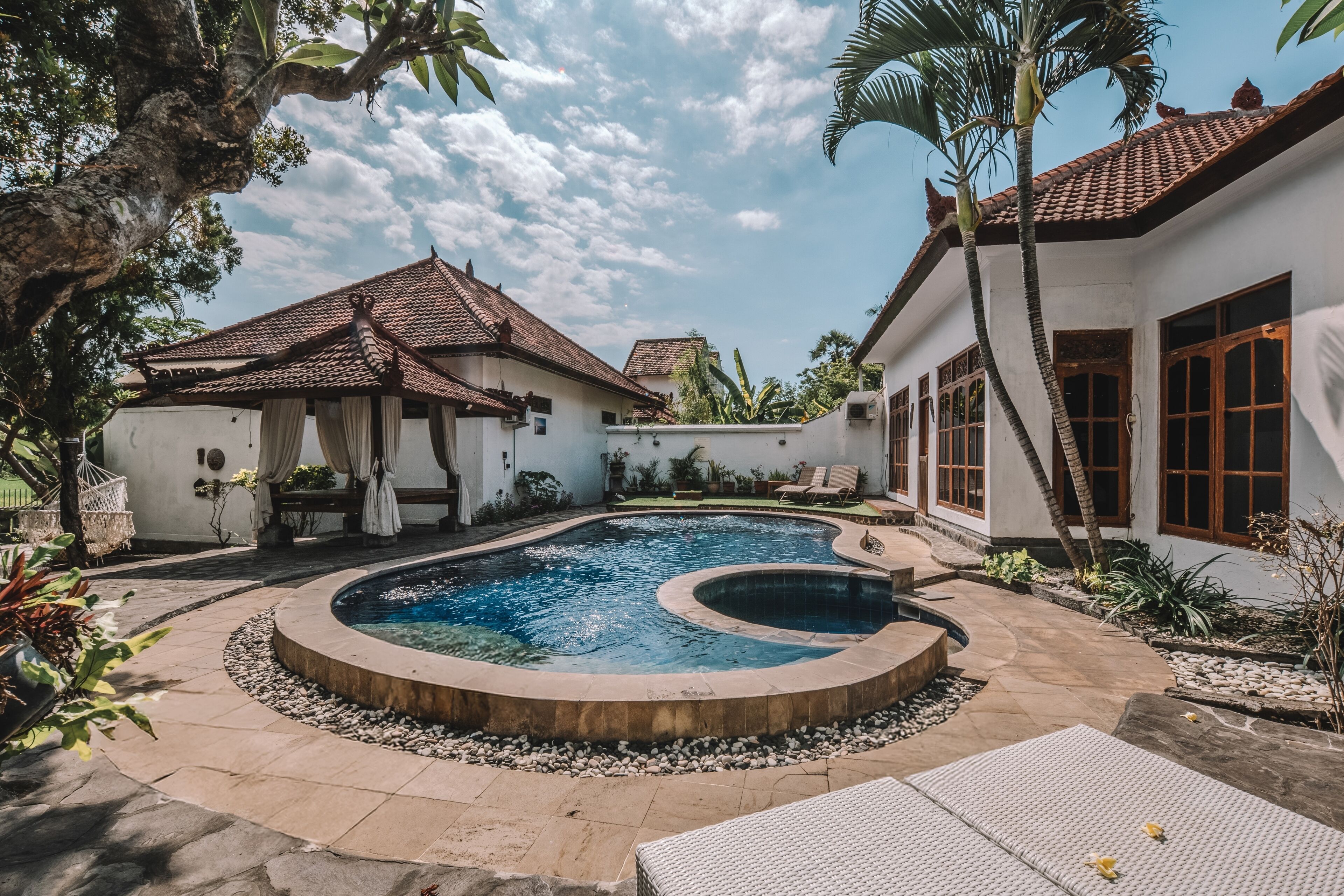 Una piscina de diseño curvo, rodeada por una terraza con borde de guijarros, y un edificio con tejado de azulejos y fachada blanca al fondo.