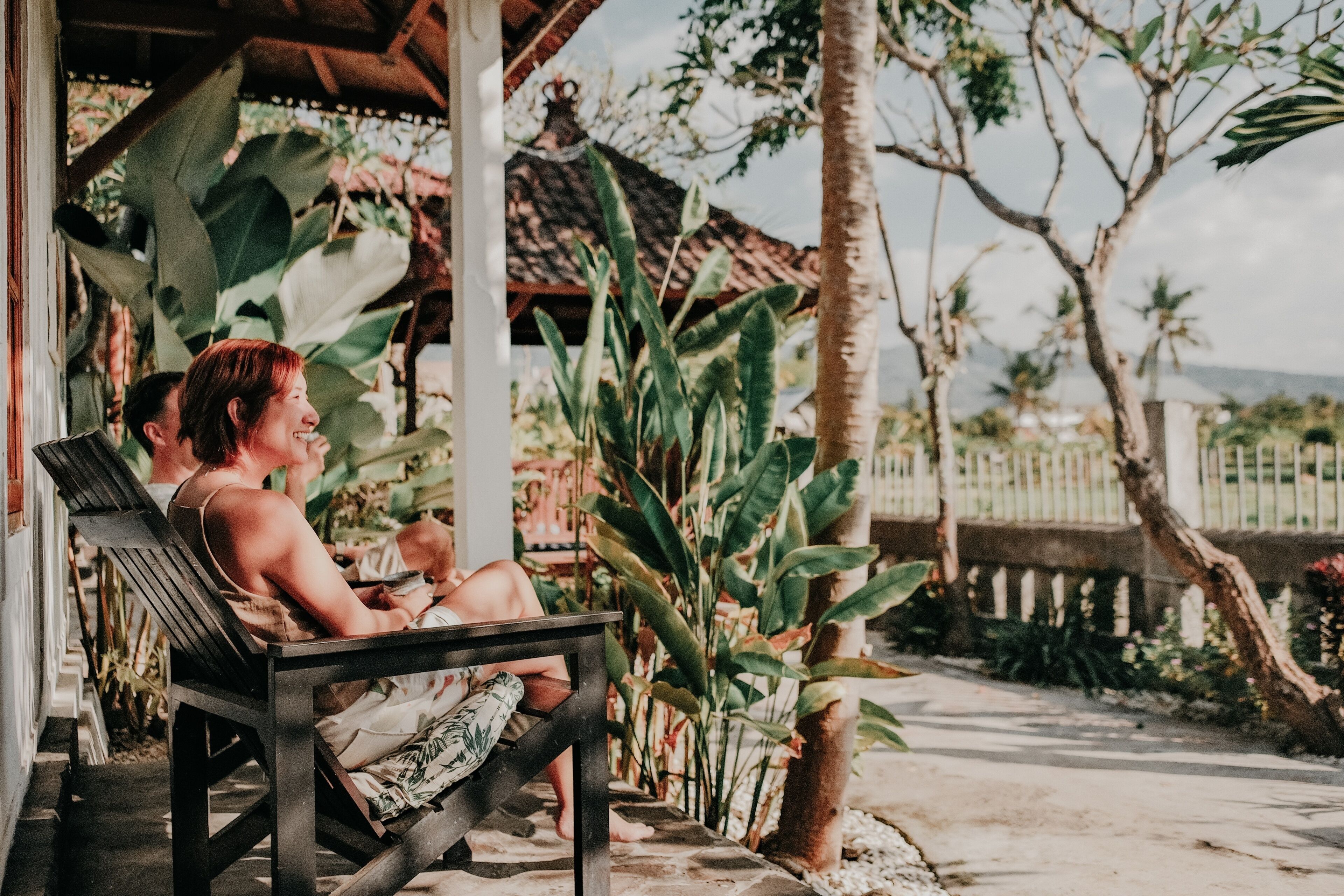 Un espacio de estar al aire libre cubierto, con un banco, rodeado de plantas tropicales y vistas a una cerca y árboles.
