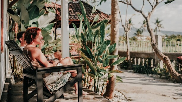 Un espacio de estar al aire libre cubierto, con un banco, rodeado de plantas tropicales y vistas a una cerca y árboles.