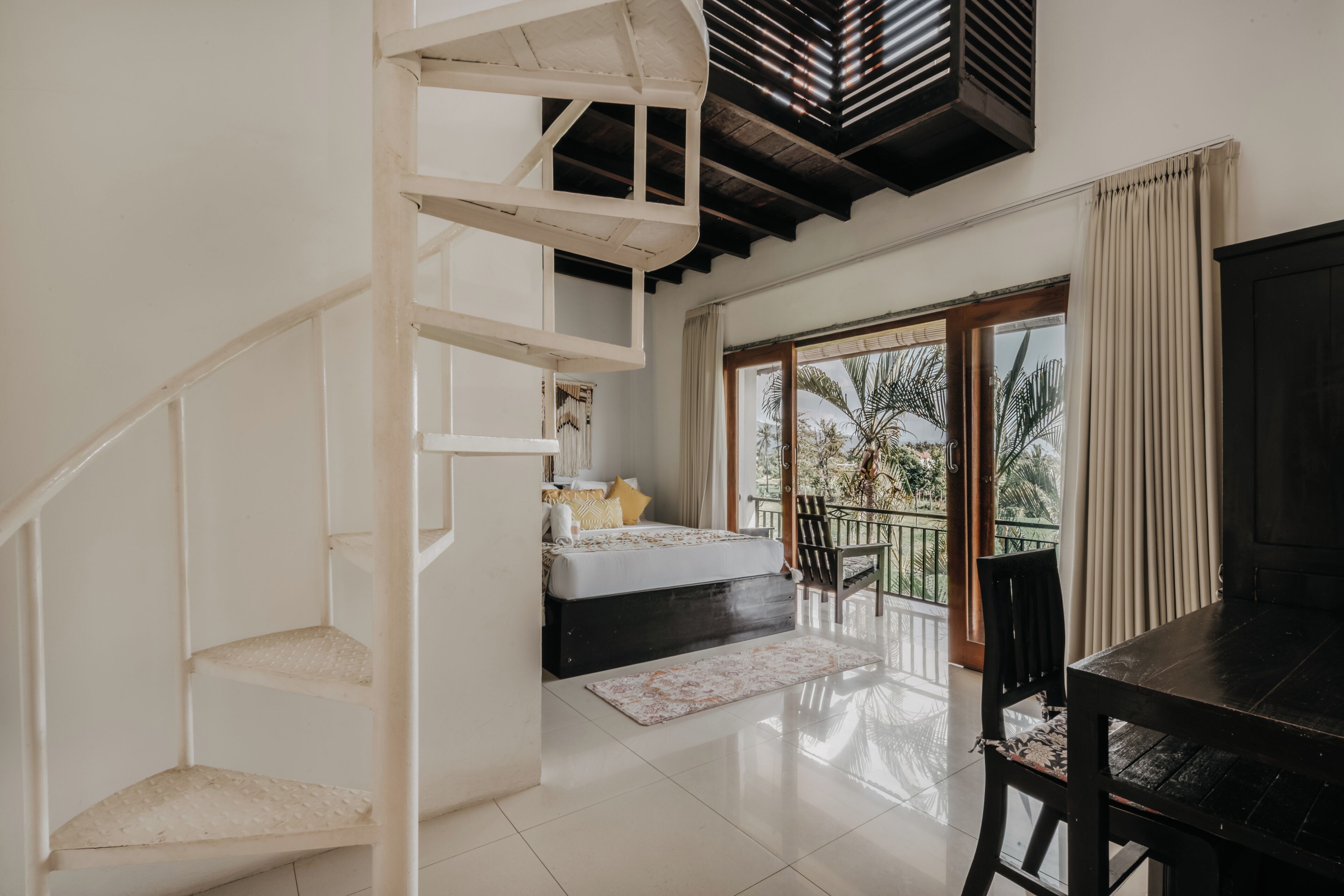Un dormitorio con cama, un escritorio pequeño, una silla, una escalera y un balcón con vistas a palmeras.