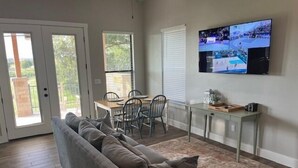 Een woonkamer met een grijze bank, een eettafel met stoelen, een aan de muur bevestigde televisie en een schuifdeur naar een buitenruimte.