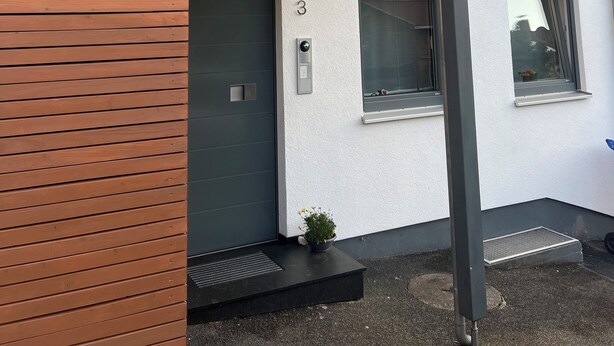 A modern two-story building with a grey facade, a wooden slatted wall on the left, and a black door with a small potted plant on the ground floor.