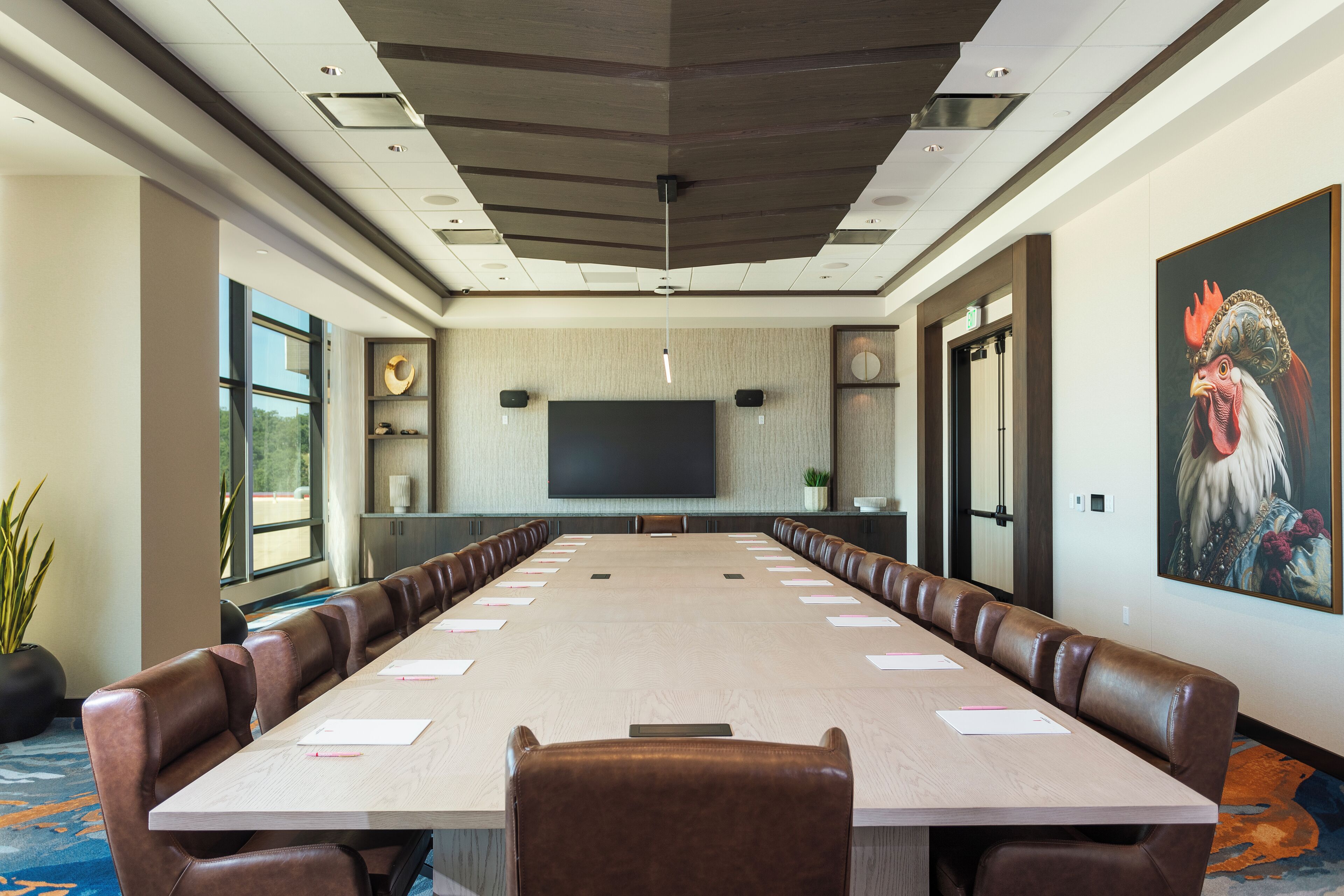 A conference room with a long table, leather chairs, a large flat-screen TV, and a painting of a rooster on the wall.