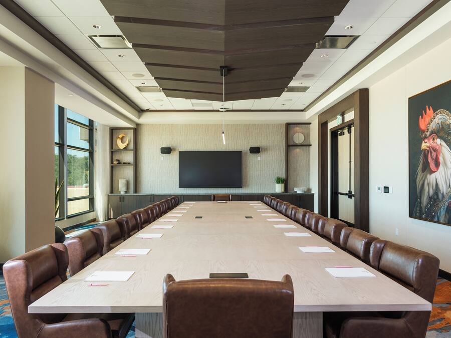 A conference room with a long table, leather chairs, a large flat-screen TV, and a painting of a rooster on the wall.