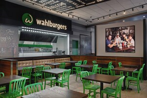 Interior de restaurante con sillas y mesas verdes, una barra con un letrero de 'Wahlburgers', y una fotografía grande en la pared.