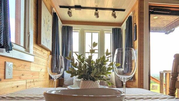 A dining table with a bowl and two wine glasses, a potted plant, and a view of the outdoors through large windows. - Charming Tiny House with Breath-Taking Views in Delano, TN (Delano)