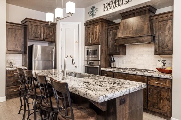 A kitchen with dark wooden cabinets, stainless steel appliances, a marble island with bar stools, and a 'FAMILY' sign above the cabinets.