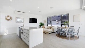 A modern kitchen with a central island, a dining area with a round table and chairs, and a living area with a sofa and a TV. - Oceanviews on Pearl (Kingscliff)