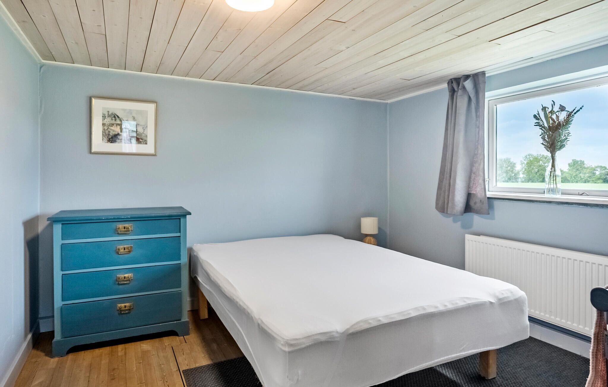A bedroom with a single bed, a blue dresser with drawers, a framed picture on the wall, a window with a view of greenery, and a radiator.