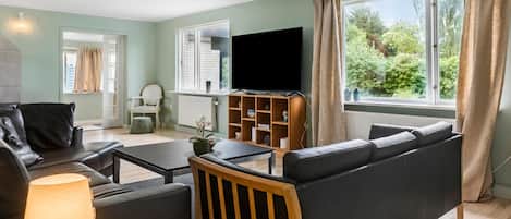 A modern living room with a sofa, a coffee table, a TV, and a bookshelf. There is a window with curtains and a view of greenery outside.