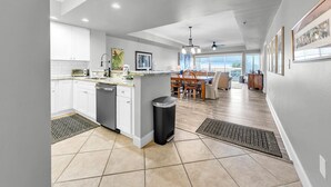 A modern kitchen with white cabinets, a dishwasher, and a dining area with wooden chairs and a table. - Harbor Landing 102B- The heart of Destin: shopping & restaurants! Beach access w/ pools & hut tubs! (Destin)