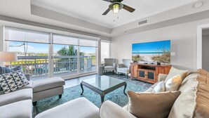 A spacious living room with a balcony, a large window, and a ceiling fan. - Harbor Landing 102B- The heart of Destin: shopping & restaurants! Beach access w/ pools & hut tubs! (Destin)