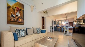 A modern living room with a beige sofa, patterned cushions, and a wooden coffee table. A painting of a woman playing a drum is on the wall. The kitchen is visible in the background with wooden cabinets and a dining area.