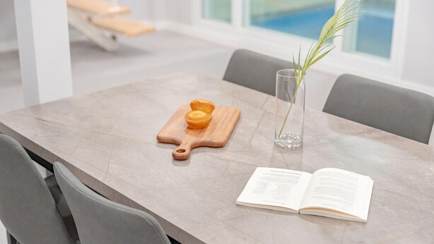 Een eettafel met een boek, een houten plank met gebak, en een glazen vaas met een plant.