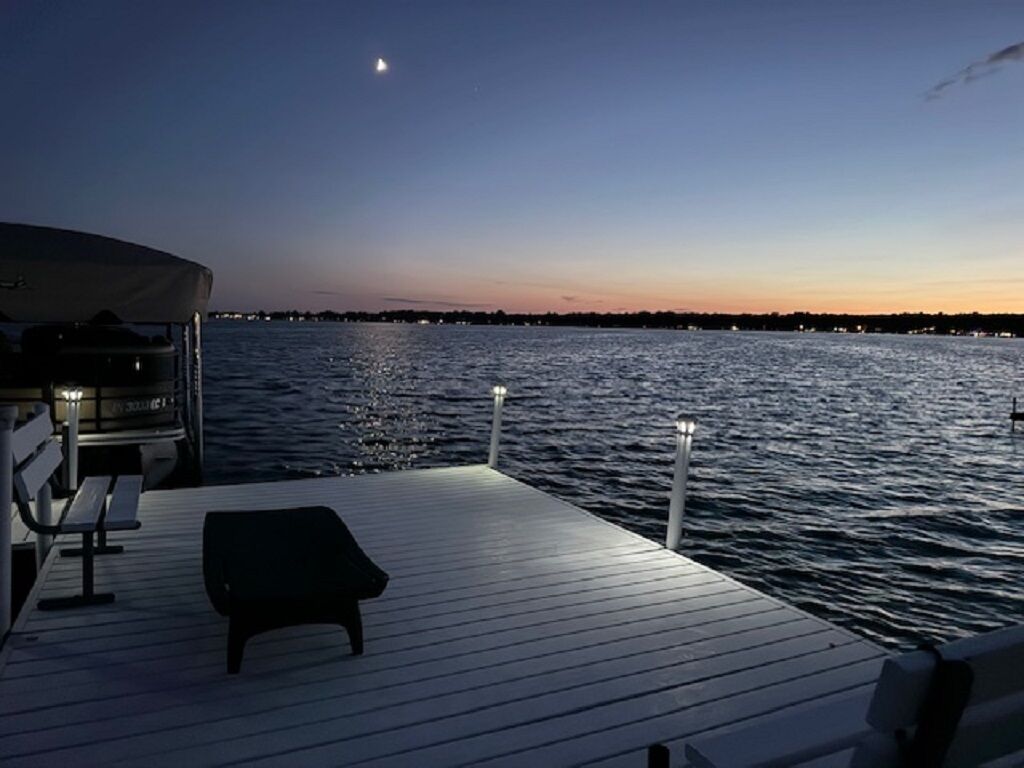 Eine Holzterrasse mit einer Bank und einem Sonnensegel, mit Blick auf ein Gewässer im Abendlicht.