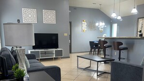 Un saló modern amb televisió de pantalla plana, sofàs grisos, una taula de centre i una zona de menjador amb cadires i una barra.