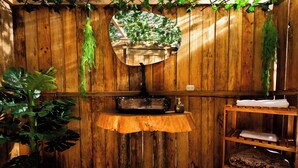 A rustic bathroom with a wooden wall, a round mirror, a small sink, a shelf with towels, and a potted plant. - Look & Luck Glamping by David (Calacali)