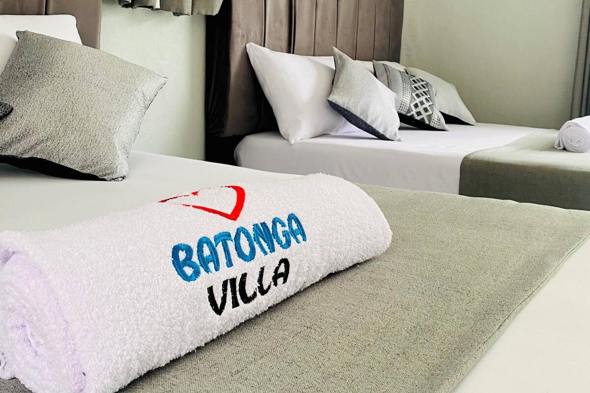 A hotel room with two beds, a headboard, and a neatly folded towel with "Batonga Villa" embroidered on it.