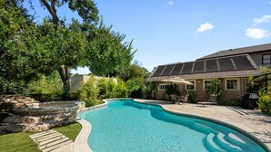 A swimming pool with a stone border, a patio area with chairs, and a house with solar panels. - Majestic Hyde Park Historic Home (Austin)