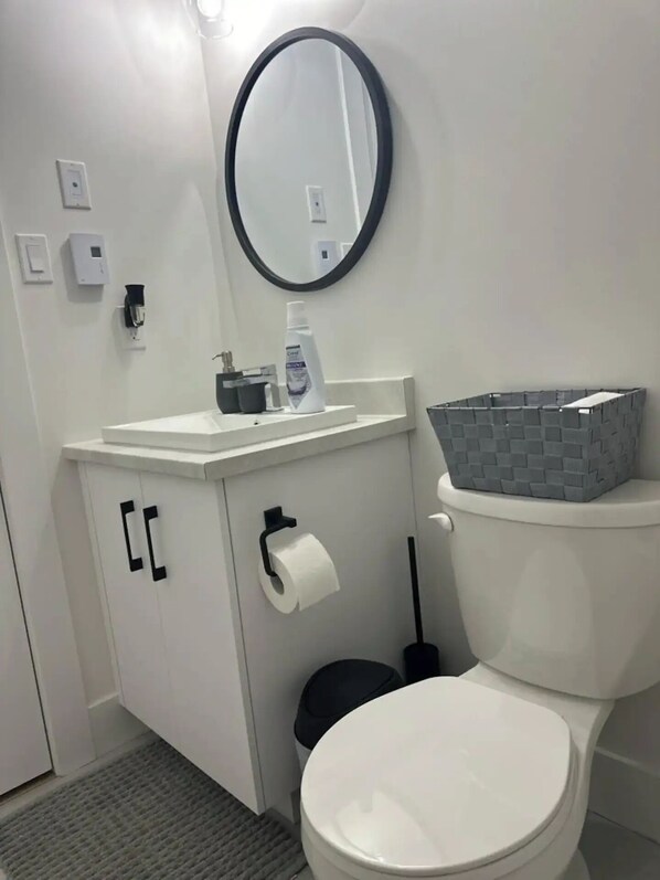 A bathroom with a white toilet, a sink with a mirror above it, a toilet paper holder, a trash bin, and a storage basket on the toilet tank.