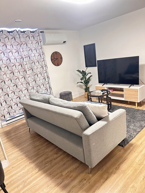 A modern living room with a grey sofa, a flat-screen TV, a wooden clock, and a potted plant. - Tynnah’s Place (Riverview)