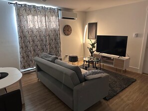 A modern living room with a grey sofa, a flat-screen TV, a round coffee table, and a decorative plant. - Tynnah’s Place (Riverview)