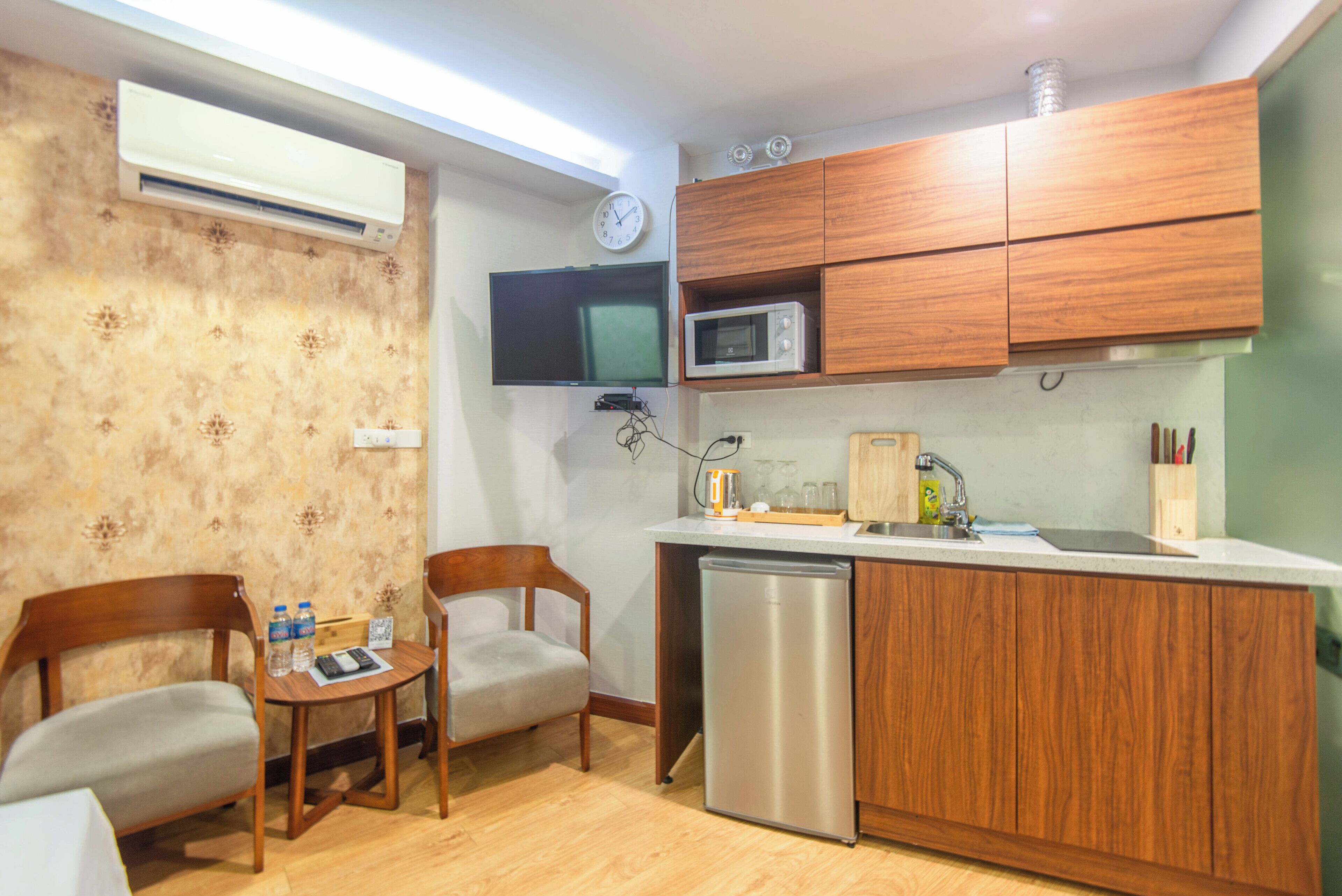 Una cucina compatta con mobili in legno, un forno a microonde, un lavandino e un frigorifero. C'è un piccolo tavolo con sedie e un orologio appeso al muro.
