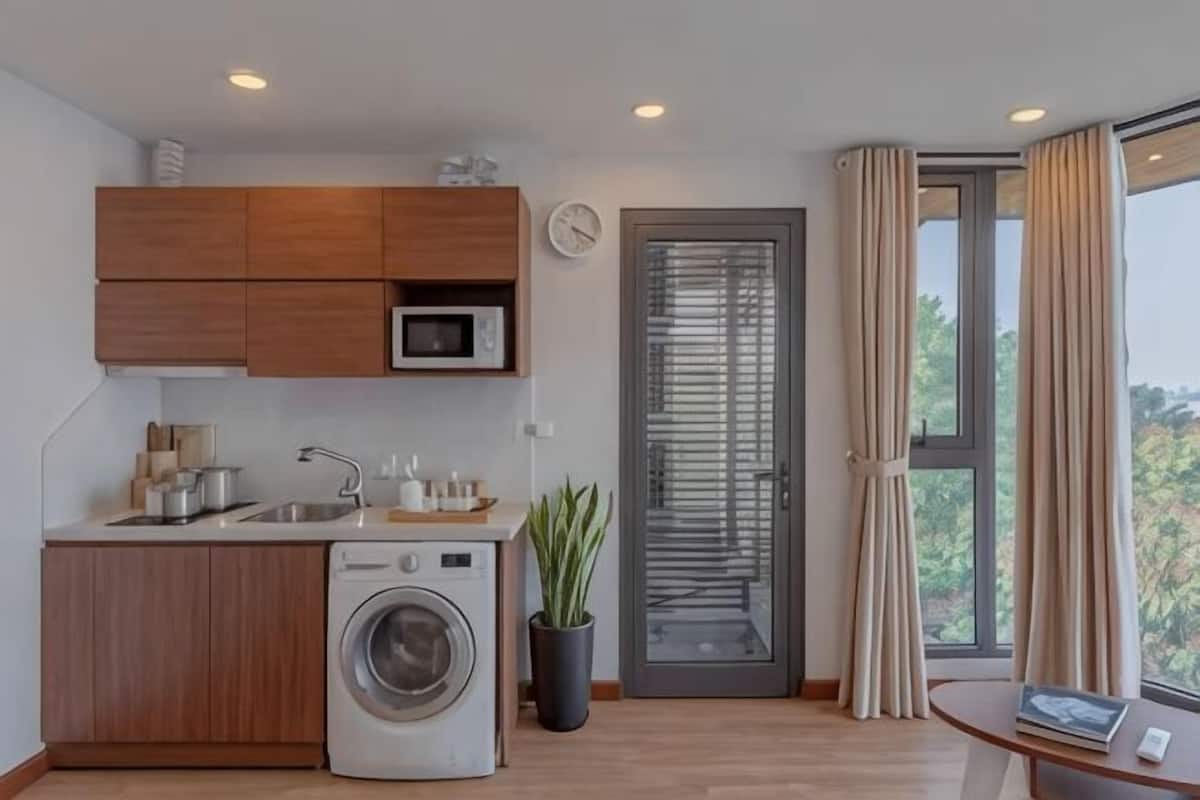 A compact kitchen with a washing machine, wooden cabinets, a microwave, and a window with curtains.