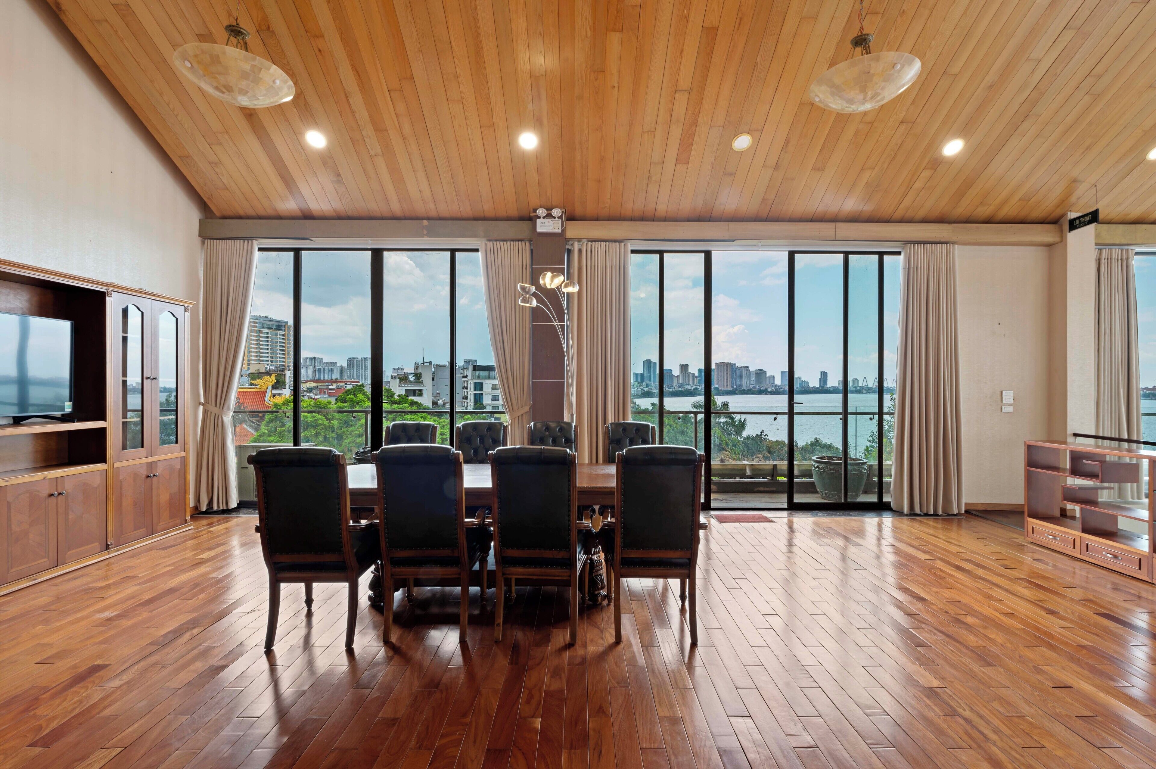 A spacious room with a wooden ceiling, large windows with curtains, a dining table with chairs, and a television on the left.